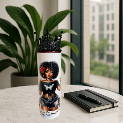 Tumbler with a design of a woman with an afro, wearing a crown, and a butterfly on her chest, placed on a surface with a plant and notebook in the background.