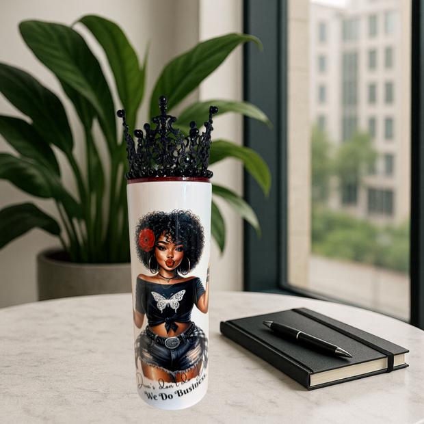 Tumbler with a design of a woman with an afro, wearing a crown, and a butterfly on her chest, placed on a surface with a plant and notebook in the background.